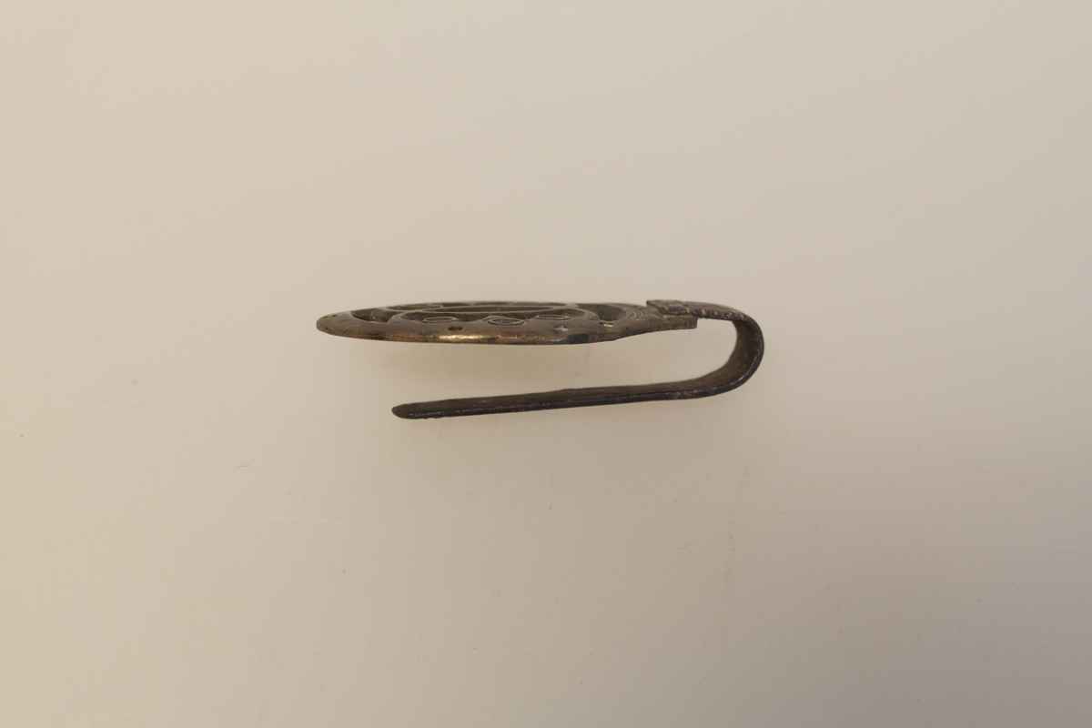 Lommekrok i støpt messing og jern. Gjennombrutt ornament i messingplaten. Kroken har huller, slik at den kan sys fast til lommen.