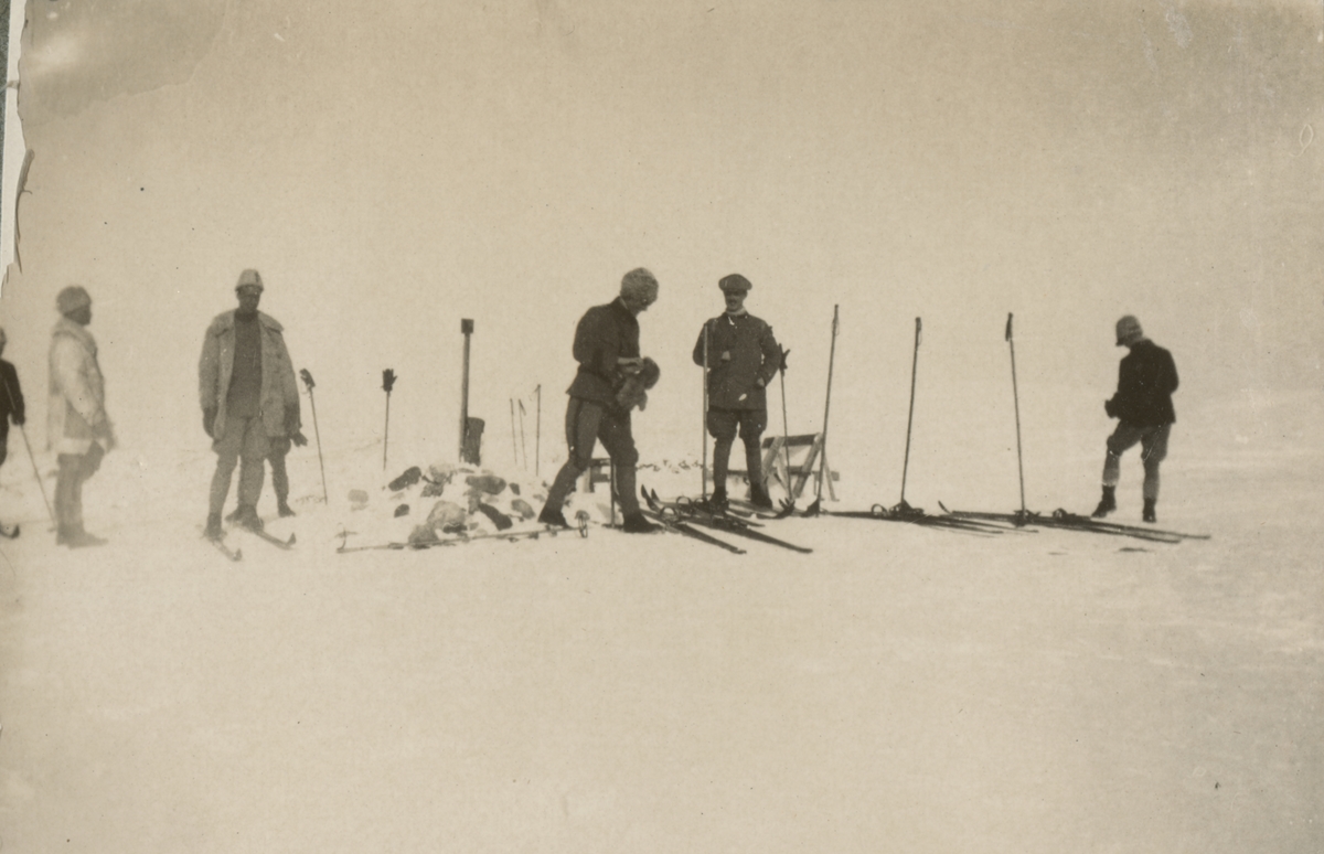 En grupp soldater vid ett stenröse på fjället. Sidor ligger på marken och stavar är stuckna i snön.