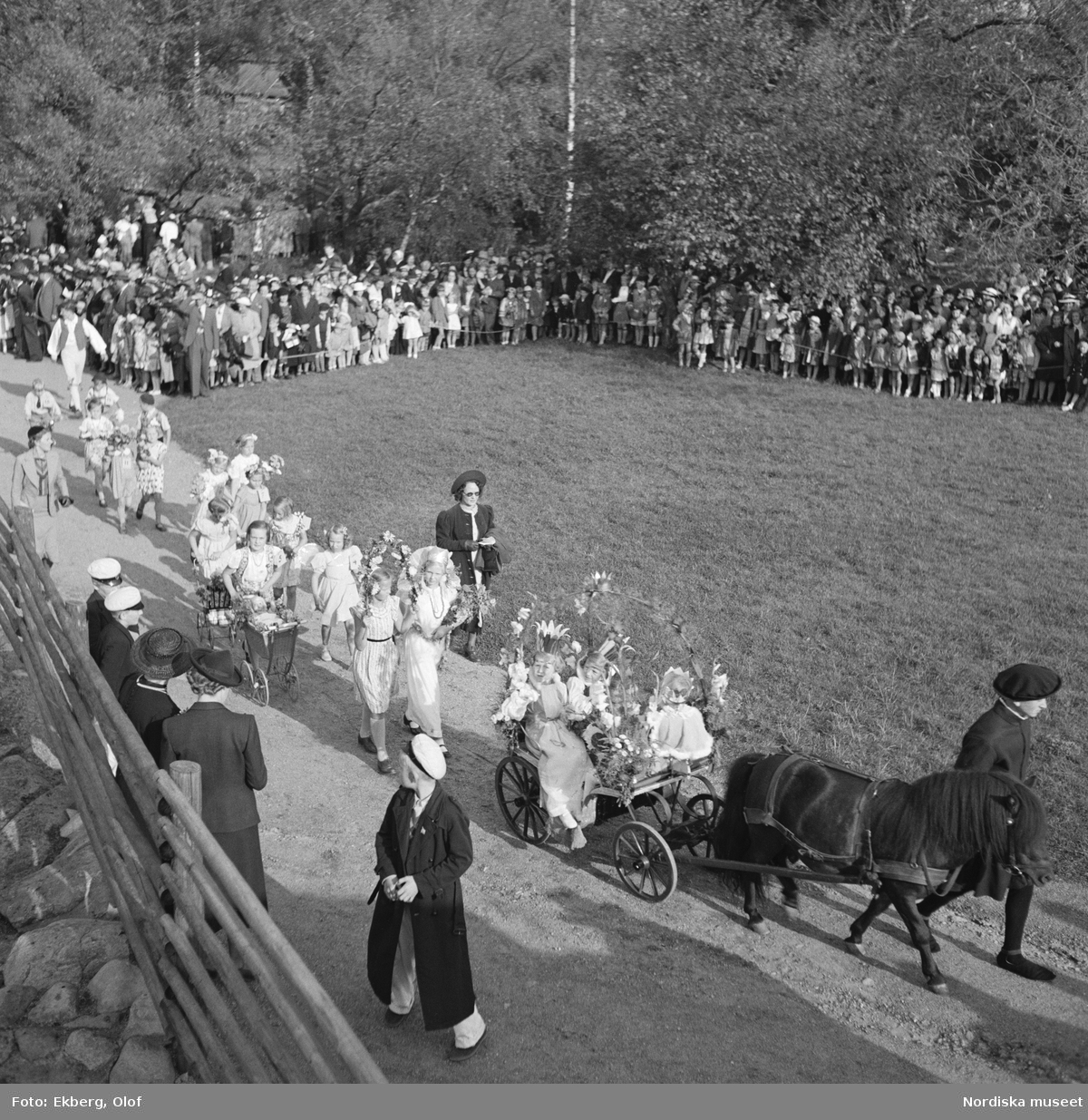 Skansen Barnens dag 1938. Bild över festplatsen tagit en bit uppifrån. Procession med blomsterprydd vagn dragen av hästar och gående parad.
