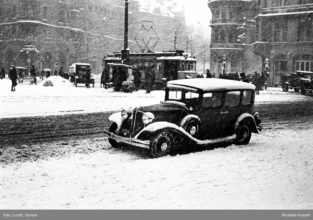 En Chrysler 1931 parkerad vid Stureplan, Stockholm. Snöfall och snö på marken. I bakgrunden fotgängare, bilar och en spårvagn.