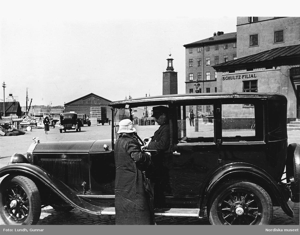 En kvinna talar med en chaufför som sitter i en parkerad Buick 1928, Klara Mälarstrand, Stockholm.