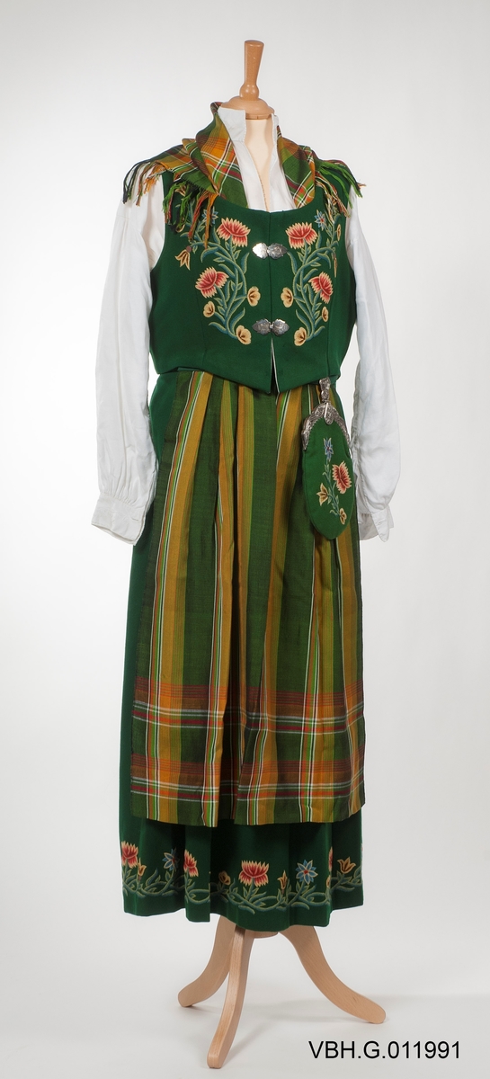 Broderier med blomstermotiver. Sjal og forkle med rutemønster.
Kjolen har blitt sydd ut i senere år med grønt bakstykke på 7 cm på hver side.