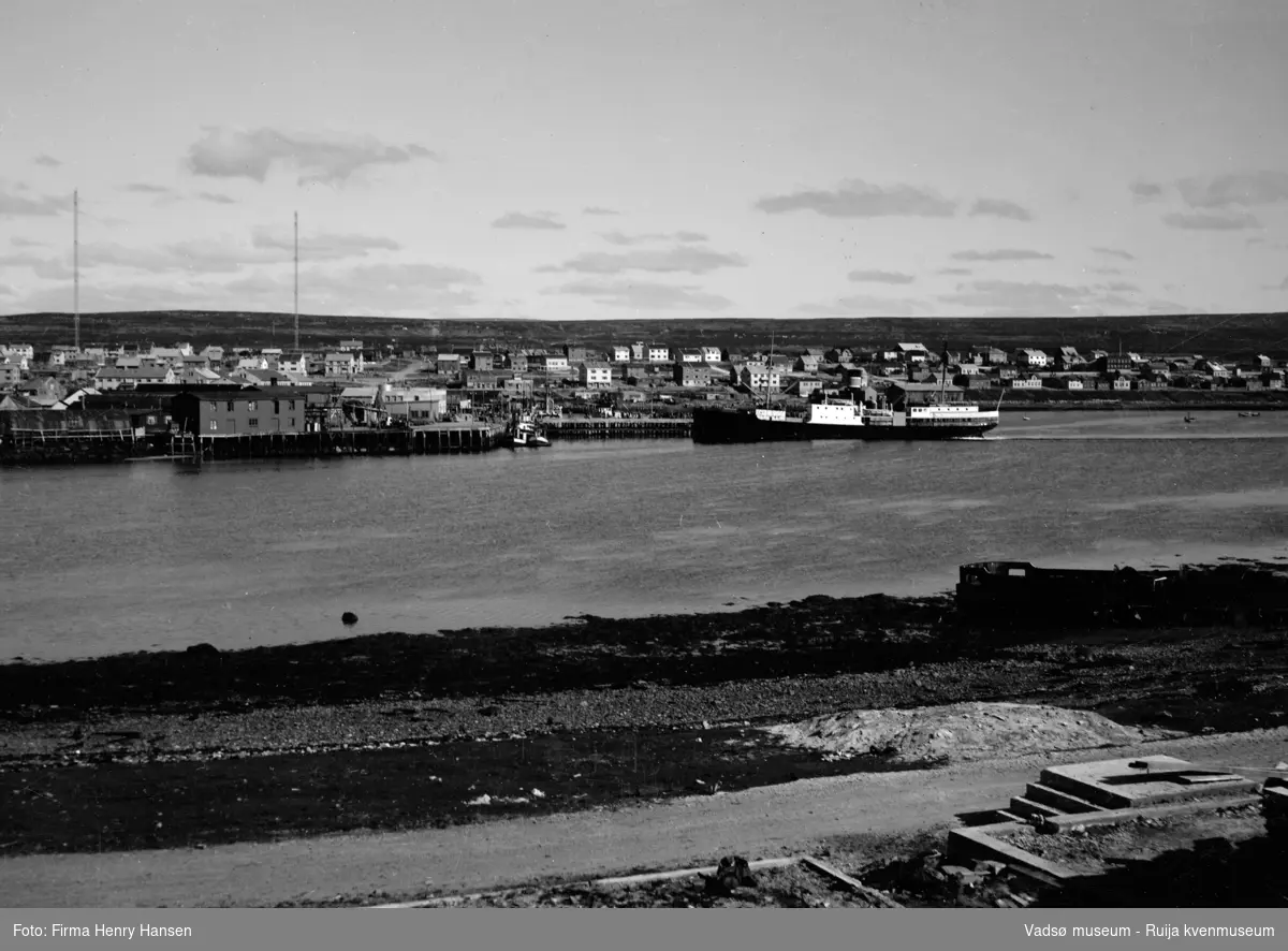 Vadsø havn. Hurtigruten D/S Finnmarken (eid av rederiet Vesteraalens Dampskipsselskap) legger til ved dampskipspiren. Indrebyen med bebyggelsen langs Havnegata og radiomastene bak. Bildet tatt fra Vadsøya, vest for broen.