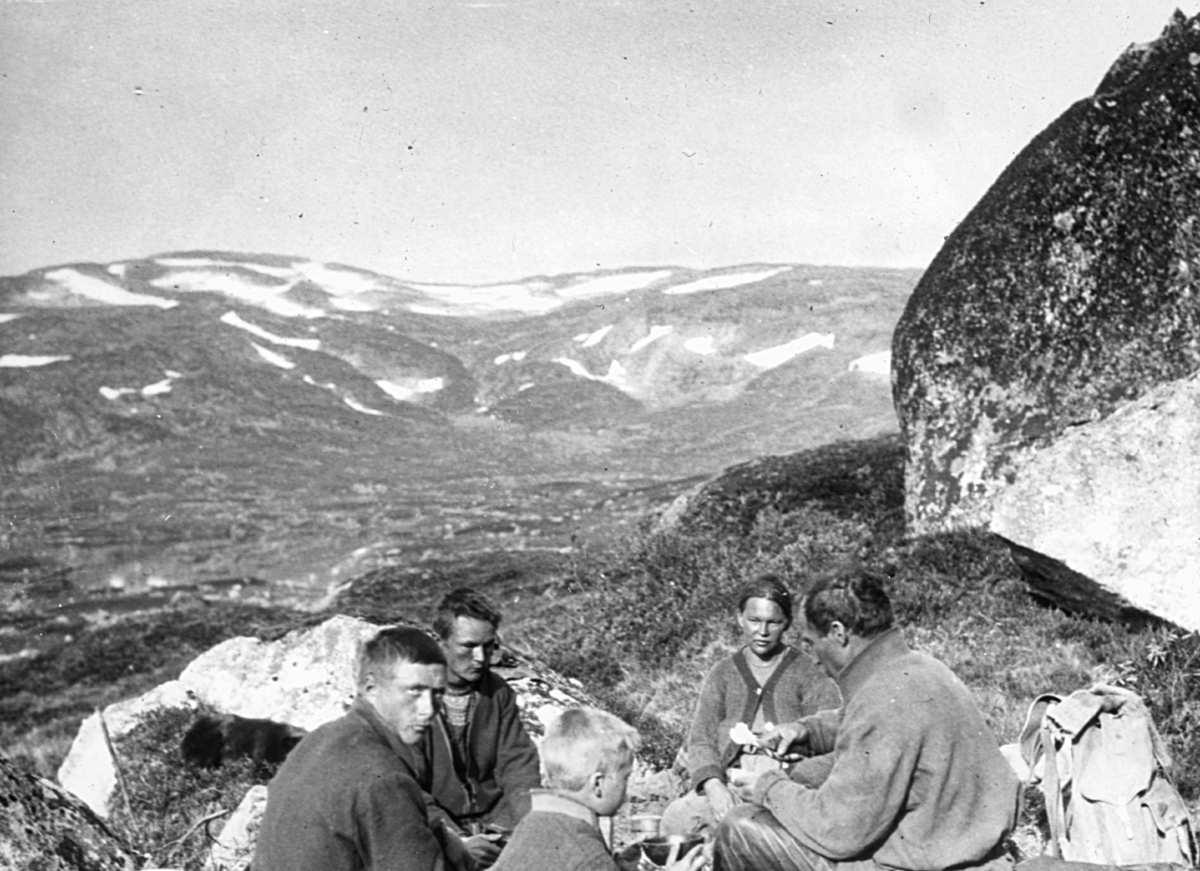 Bildet er fra Kalvvatn-området sør i Nordland. Nistepause. En gruppe samer sitter i ring og spiser. I bakgrunnen ser vi fjellandskap med snøflekker. Det er sommer og sola skinner.