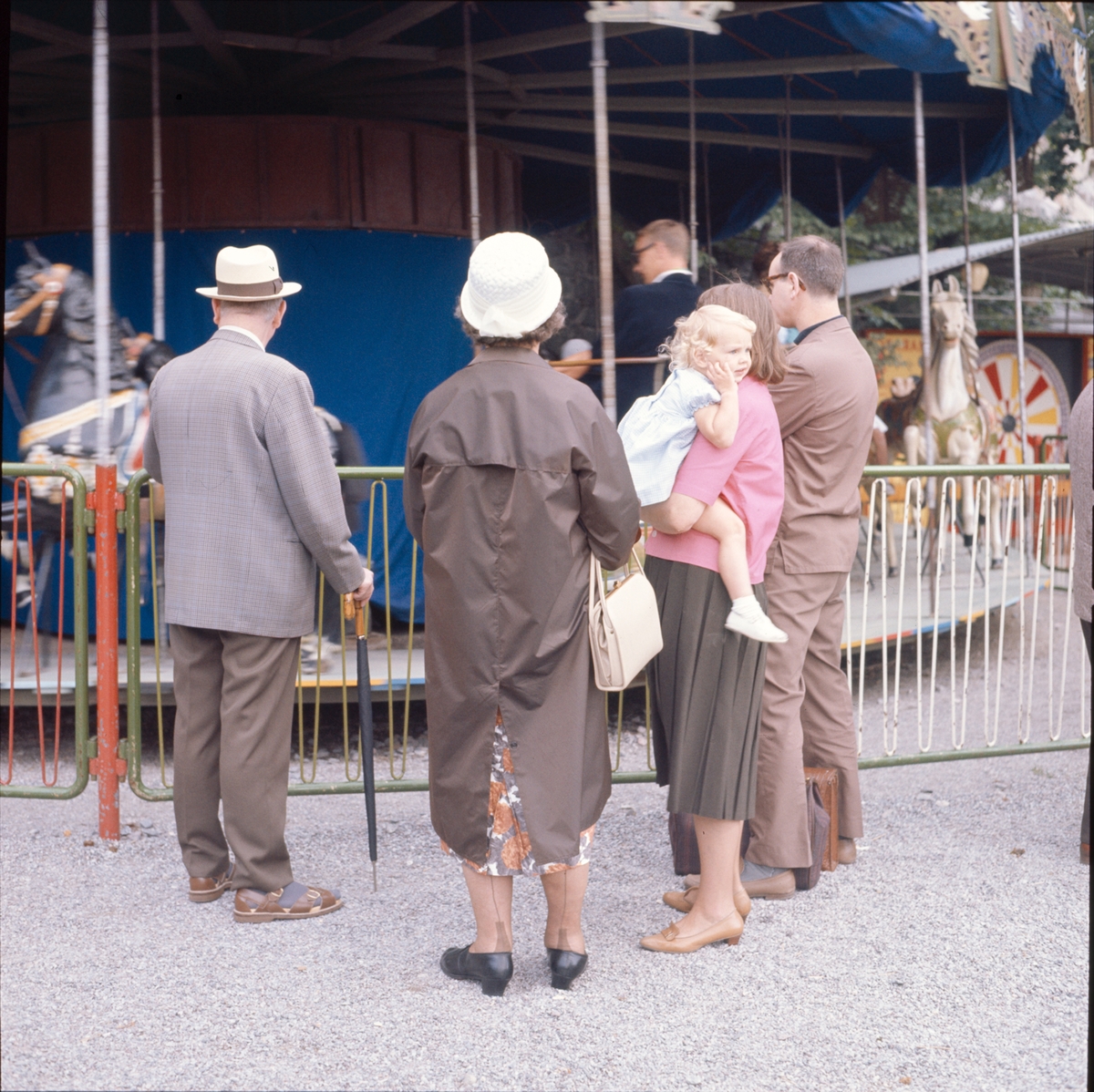 En grupp personer betraktar en karusell. Ett barn betraktar något som är mer spänande och som inträffar utanför bilden.