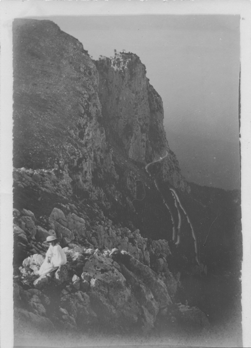 Ester Ellqvist sitter på en klippa på Capri i Italien. Den slingrande vägen i bakgrunden leder upp till Anacapri med Barbarossas borg på klippspetsen.