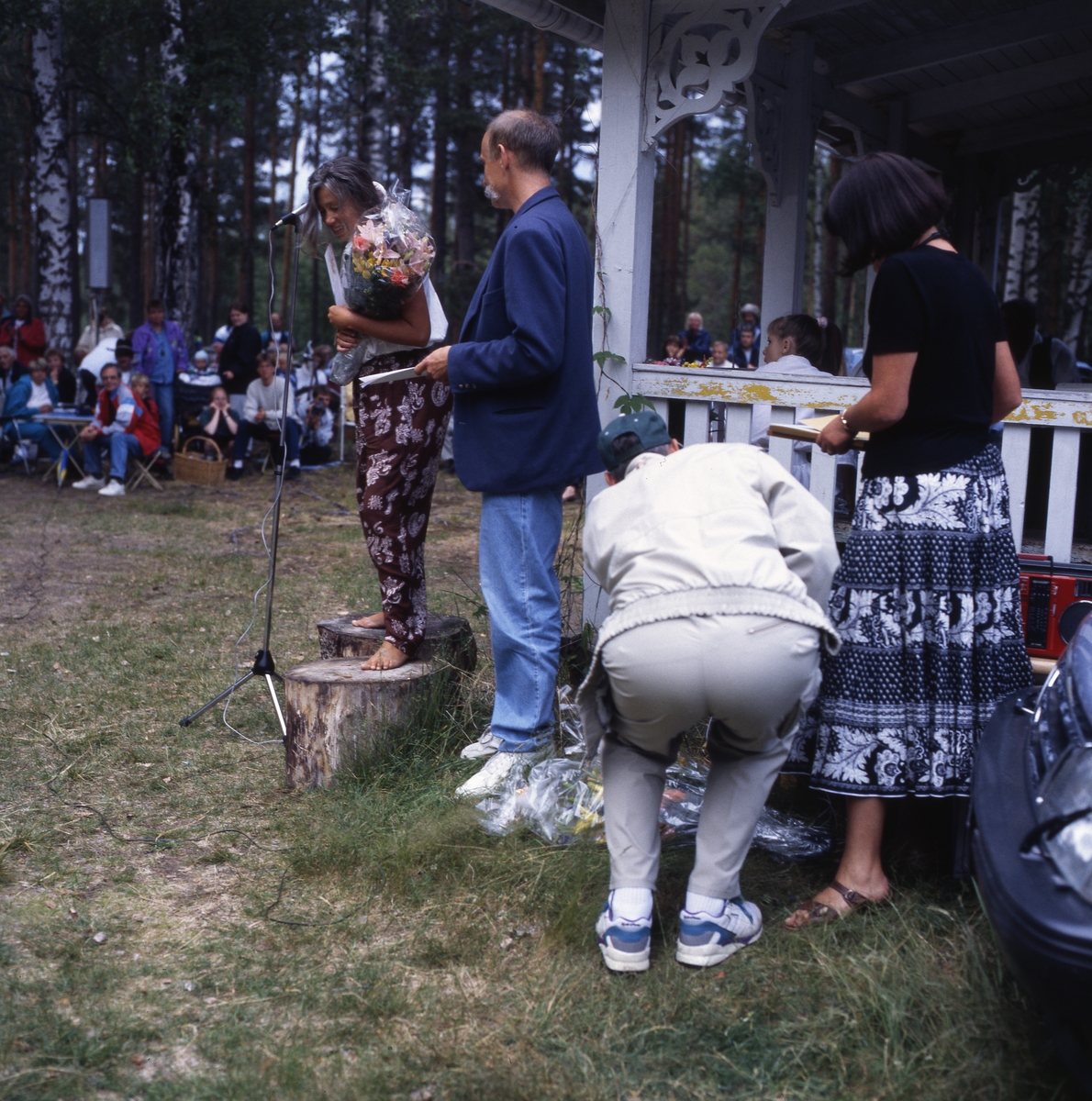 Viksten-aftonen, 11 juli 1994. En grupp männikskor sitter runt en gräsplan vid skogsbrynet. En kvinna står på en stubbe framför en mikrofon och håller en blombukett i famnen.