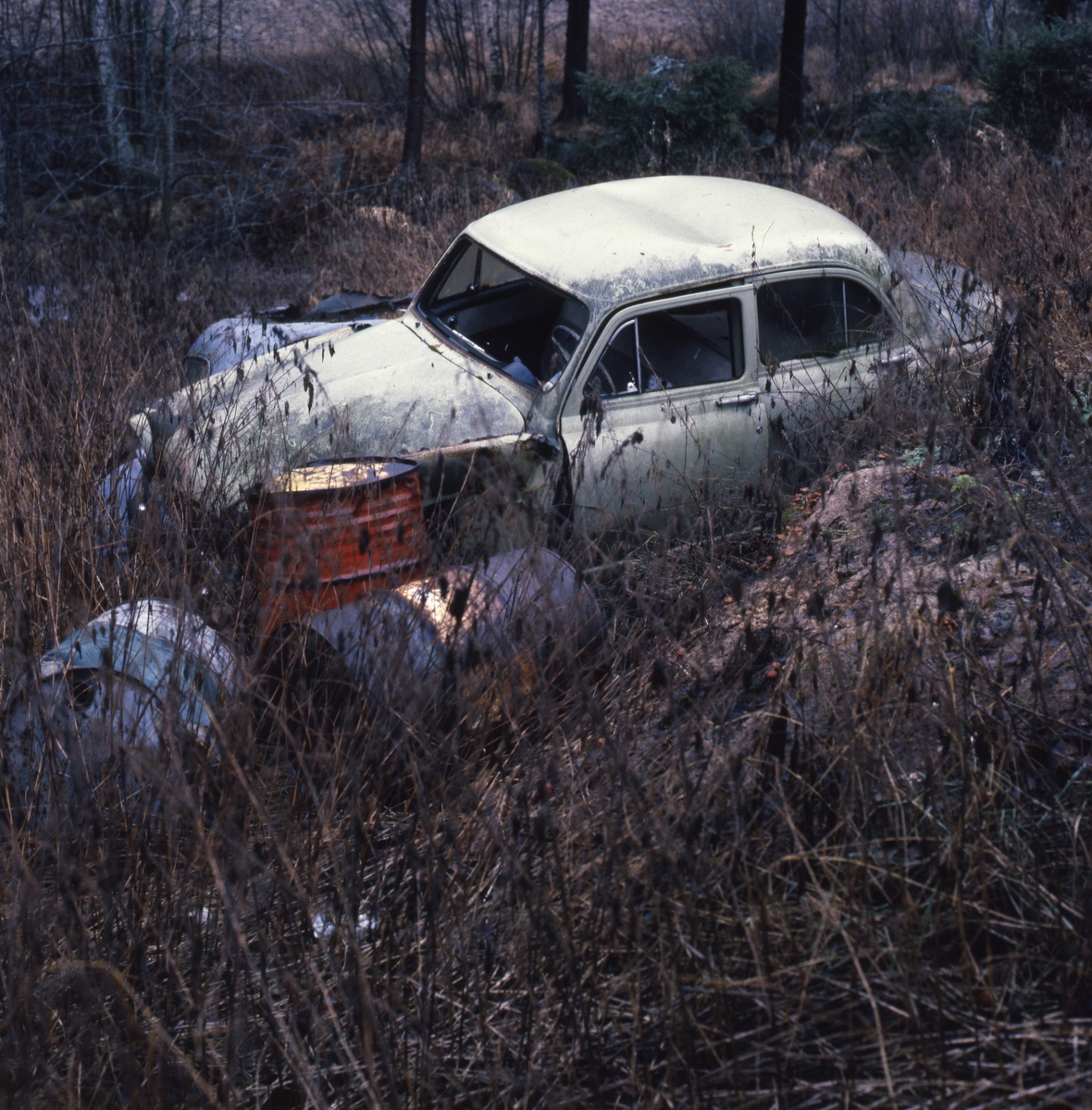 Färdigåkt, 1986. En skrotbil bland gamla rostiga oljefat och torrt ris i en slänt.