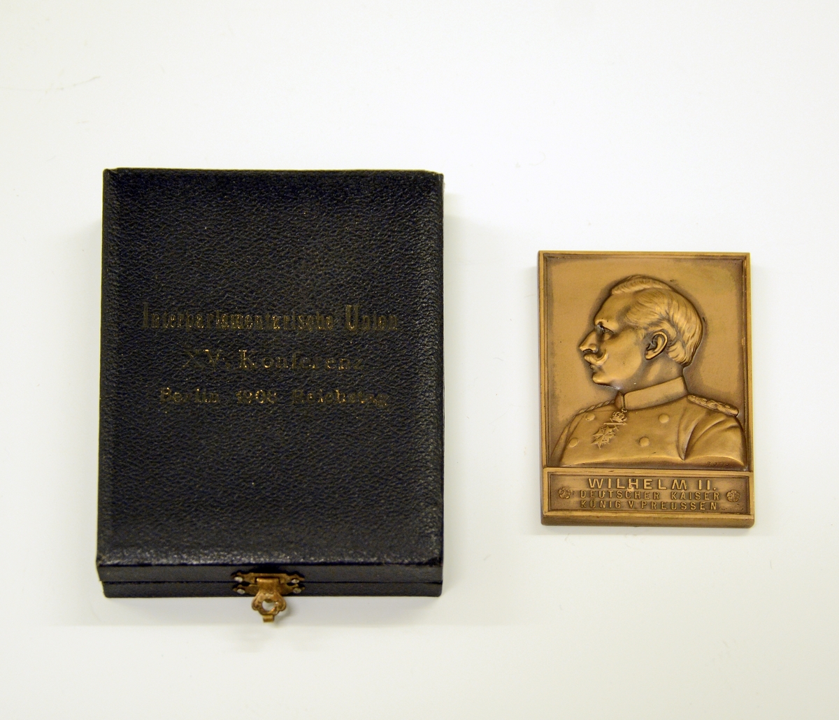 Fra protokollen: 
Plate i bronse, i etui. Platen er rektangulær, med målene 6,8 cm x 4,7 cm. På forsiden istøpt bilde av keiser Wilhelm II, hans navn og "DEUTSCHER KAISER KØNIG V. PREUSSEN" under bildet. På den andre siden bilde av en gudinne (?) som står med en globus under hvert ben, i venstre hånd en dokumentrull med ordet "PAX" på, i høyre holder hun en plantegren. Rundt figuren står følgende:   "+ 15. KONFERENTZ - DER - INTERPARLAMENTARISCHEN - UNION +". 
Ved siden av figuren: "Berlin 1908" "Reichstag".
Etuiet har sort imitert skinntrekk og på forsiden står: "Interparlamentarische Union XV. Konferentz. Berlin 1908. Reichstag". På innsiden av lokket foret med grønn silke. I gullbokstaver står: "R. OTTO. Kgl. Hofgraveur. Berlin. U. d. Linden 40." Bunnen er innvendig trukket med mørk grønn fløyel.