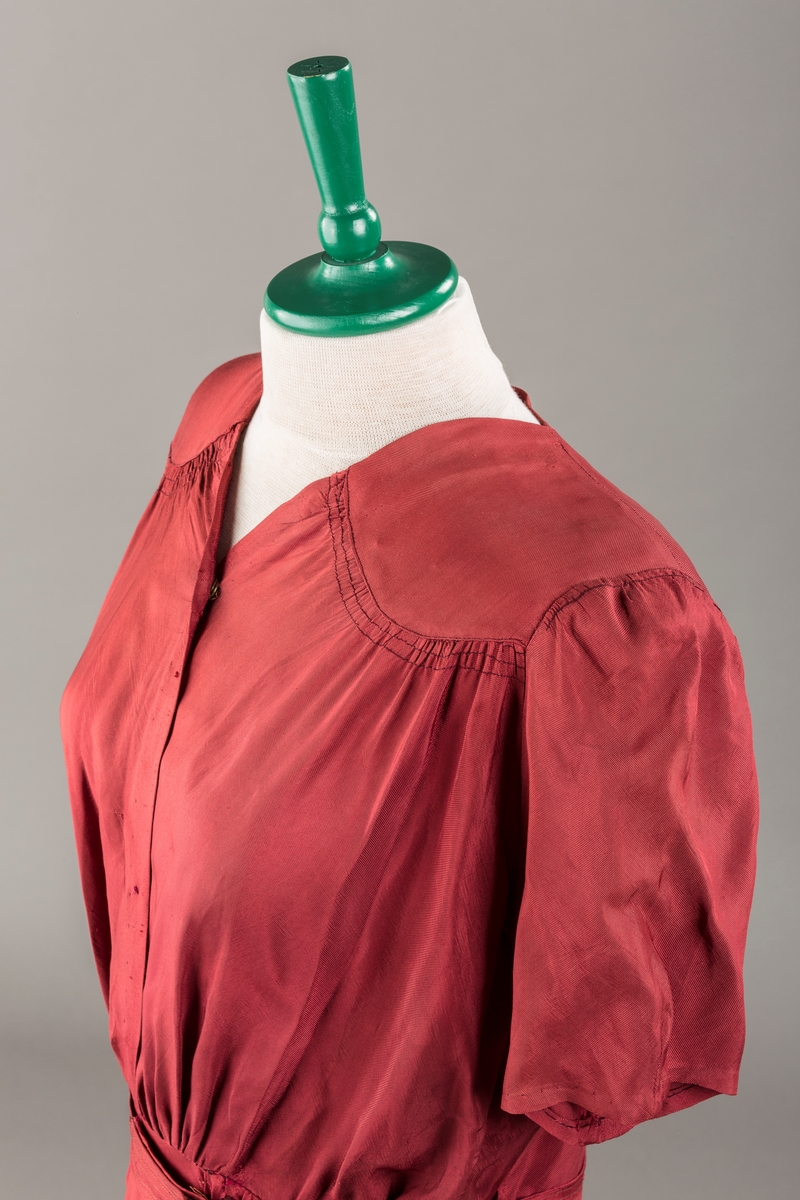 Form: Glatt, klokkeformet skjørt. Forstykkene rynket til buede bærestykker foran. Overdel rynket til skjørt. Korte ermer. Lukket med trykknapper. Belte fastsydd.