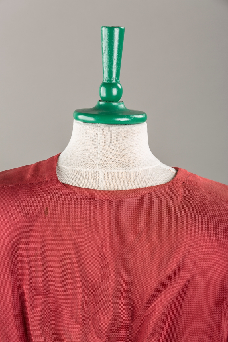 Form: Glatt, klokkeformet skjørt. Forstykkene rynket til buede bærestykker foran. Overdel rynket til skjørt. Korte ermer. Lukket med trykknapper. Belte fastsydd.