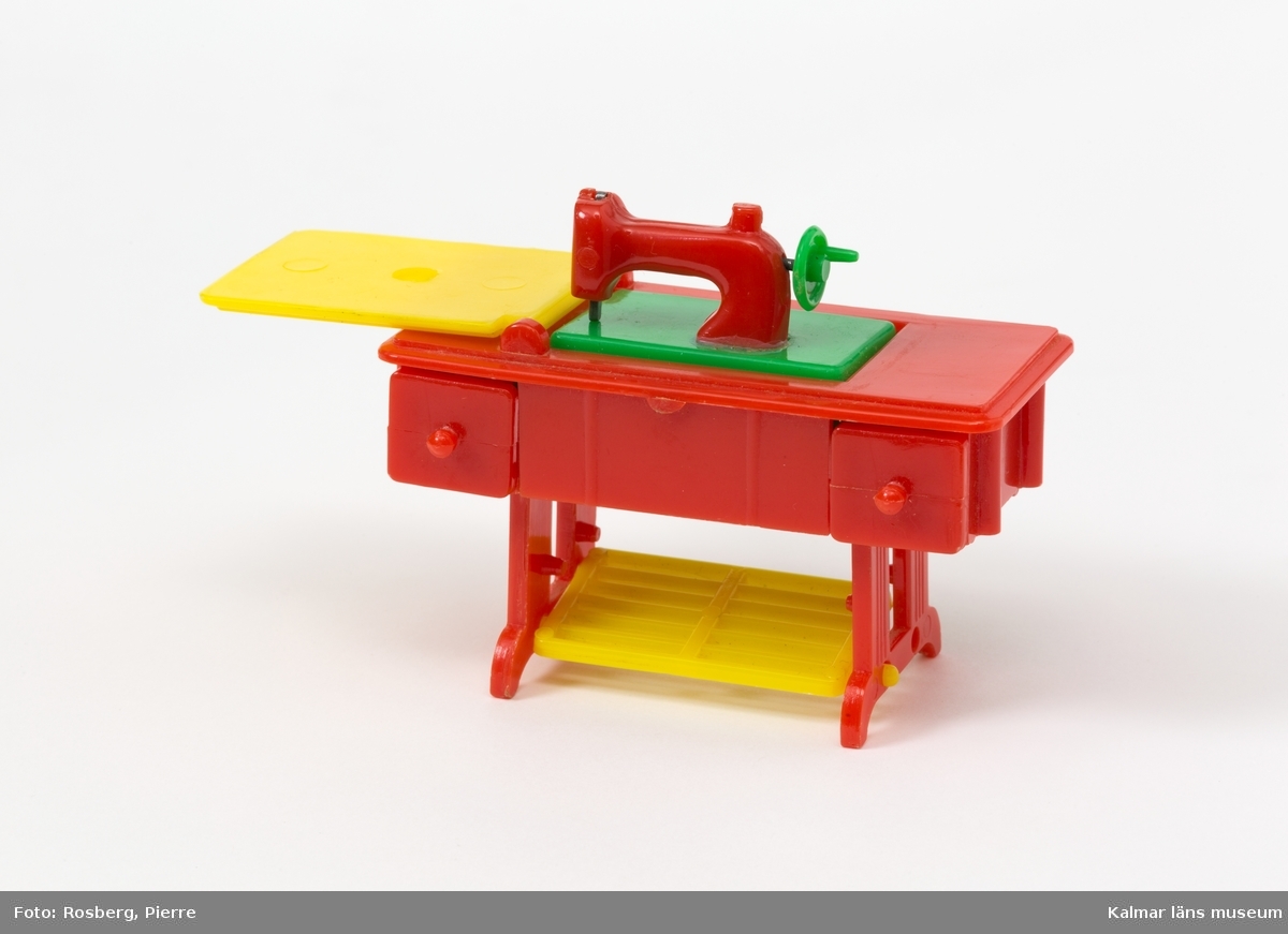 KLM 45437:7 Symaskin, av plast, dockskåpsmöbel. Bord i vilket en symaskin är nedsänkt. I det röda plastbordet finns en gul lucka som man lyfter upp och under den finns en symaskin som kan vikas upp. Symaskinen är i röd och grön plast. I bordet finns två lådor och en gul trampa/pedal.