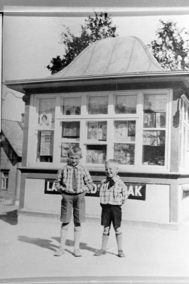 To gutter i kortbukser, fotografert foran kiosken i byskillet.