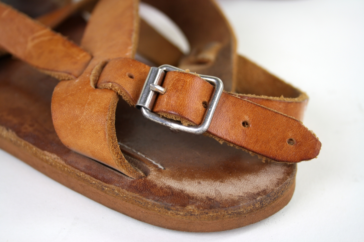 Ett par sandaler i naturfägat läder, med remmar, platt rågummisula och ett metallspänne. 
Damsandaler i storlek 37, präglat in i sulan tillsammans med tre ytterligare bokstäver eller siffror, oläsligt.