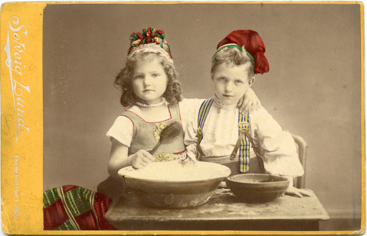 Kolorert studiofotografi av jente og gutt med drakt, sittende ved bord med to treboller, ei sleiv. 