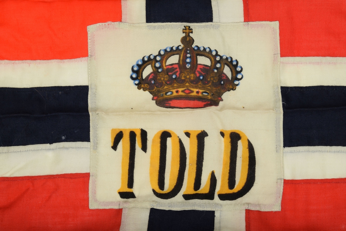 Norgesflagg med "TOLD" skrevet på midten. Over teksten er det avbildet en krone. Bånd til oppheng i den venstre kortsiden. 3 spisser i høyre side.
