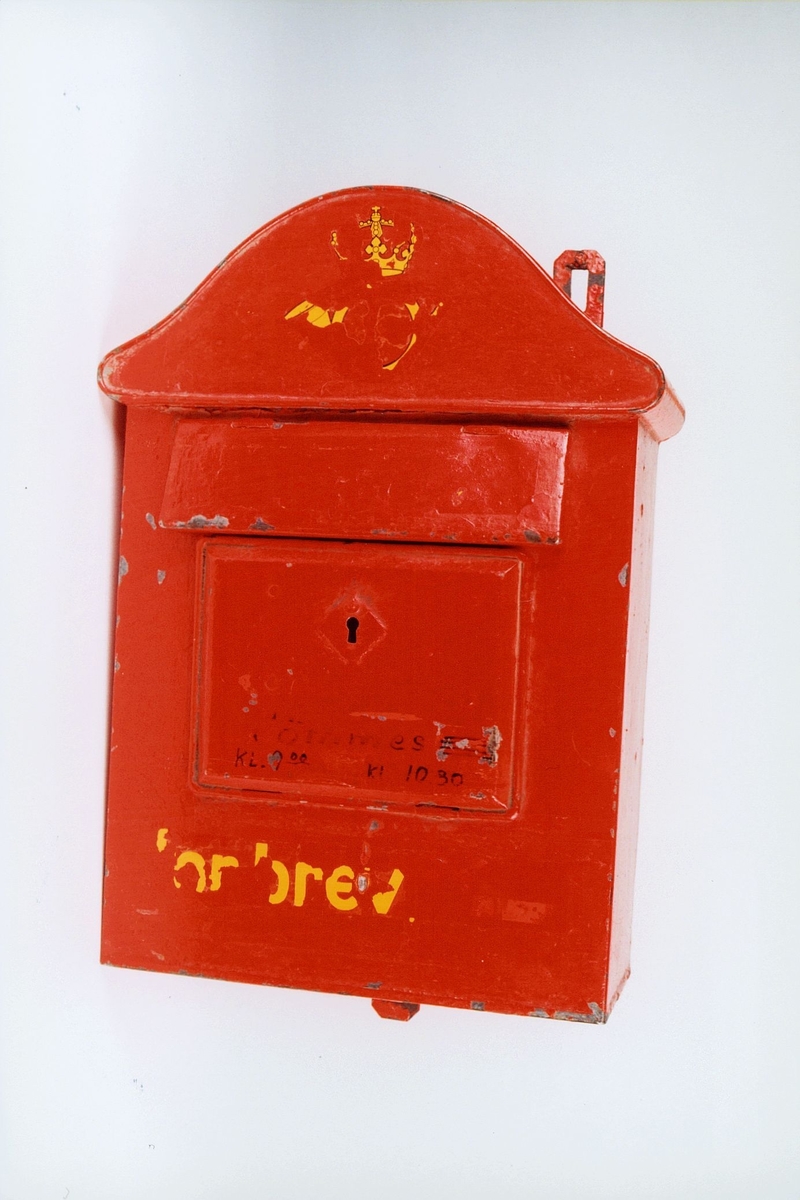 Rød postkasse med åpning og lås i front. Posthornemblem og skrift delvis nedslitt