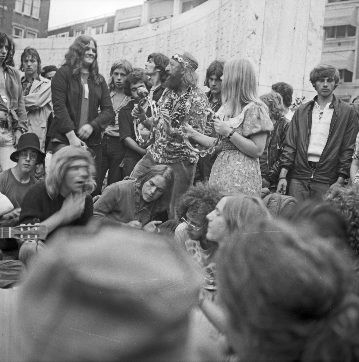 En gjeng som er samlet og spiller og synger på en åpen plass i sentrum av en by. Muligens Amsterdam.