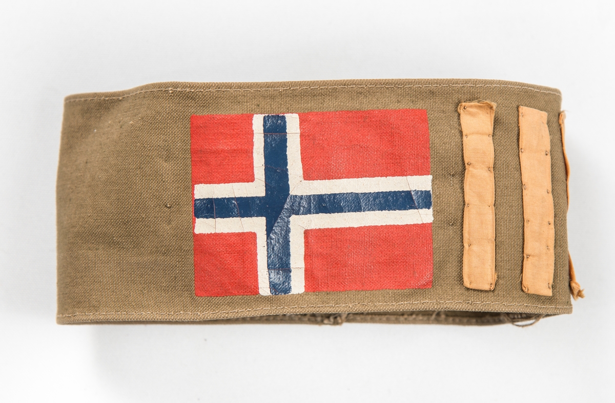Armbind (Kaptein) i brunt bomullstøy. Påtrykt det norske flagget og påsydd tre tynne gule striper. Var sett på ermet til VFF 09539 Uniformsjakke (med knappenål).