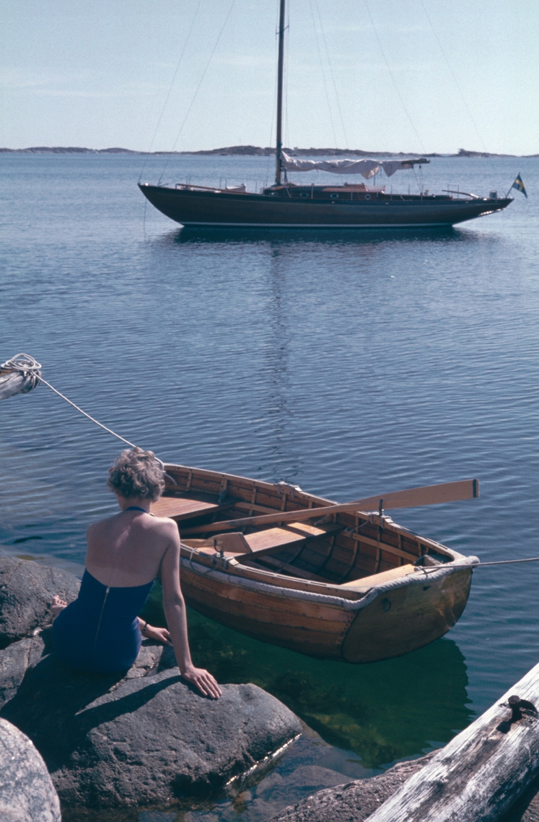 Kvinna med baddräkt på klipphäll i skärgården, segelbåten Elsi-Anne och en mindre roddbåt ligger förtöjda i närheten.