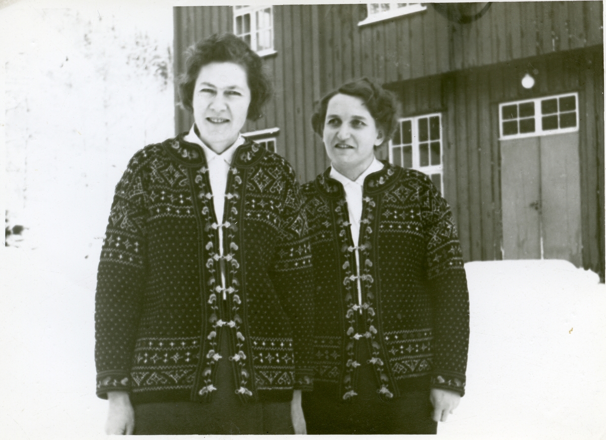 2 kvinner står utenfor et hus. Begge ha på seg hvite bluser og strikkekofter. Det er snø.