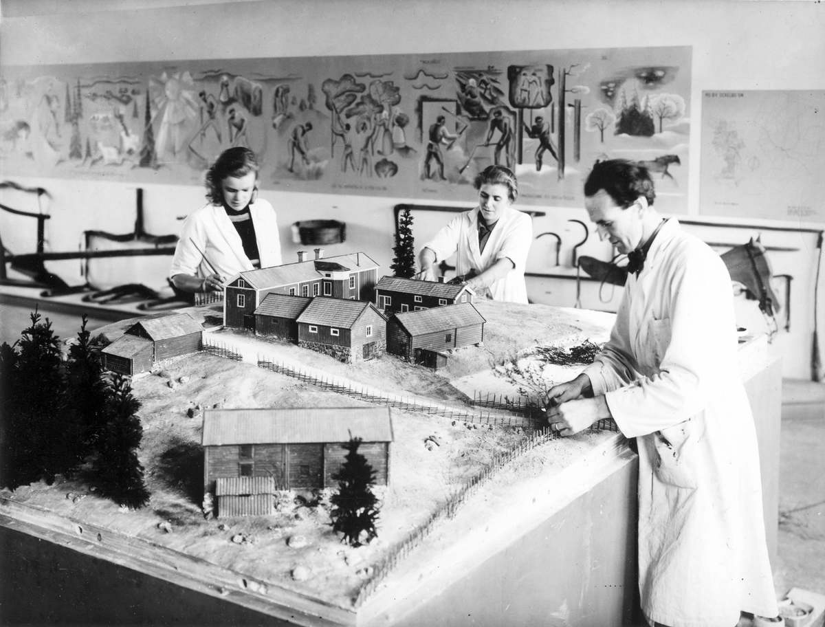 Tillverkning av modellen till gården "Västergårds". Allmänninge i Valbo 28 oktober 1941.
Mannen på fotot är konstnären och modellbyggaren Manne Östlund från Valbo. I mitten hans hustru Brita och till vänster Ulla Hägglund från Bergby i Hamrånge.