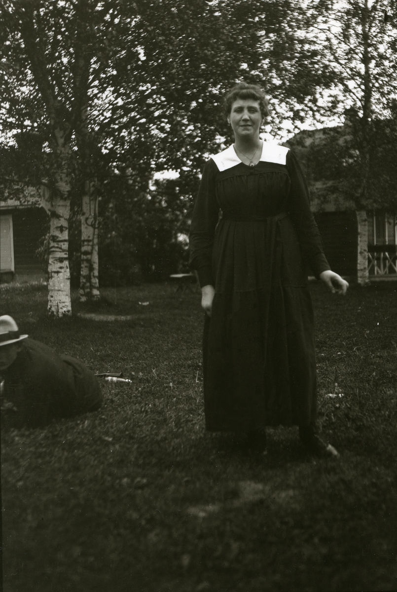Kvinne, Greta Karlson, i Malung. Hus og bjørker i bakgrunnen. Mann liggende på bakken til venstre.