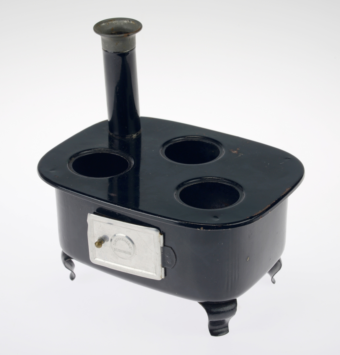 En liten komfyr med tre kasseroller og en steikepanne. Gjenstandene er undernummerert 01-05.
FTT.54454.01: En svartlakkert lekekomfyr som er firkantet med avrundede hjørner. Den er trolig laget av jernblikk og blikk. På kokeflaten er det tre hull for å plassere kasserollene på. Bakerst til venstre er det en 8cm høy pipe. I front er det en sølvfarget blikkdør som er festet med hengsler. På døren er det innskrift og et lite messingfarget og rundt håndtak. Komfyren har buede bein.
FTT.54454.02: En høy kasserolle laget av blikk. I hver side er det loddet på håndtak. 
FTT.54454.03: En lav kasserolle laget av blikk. I hver side er det loddet på håndtak.
FTT.54454.04: En lav kasserolle laget av blikk. Det har vært festet et langt håndtak i den ene siden, men det er nå løs.
FTT.54454.05: En steikepanne laget aav blikk. Håndtaket mangler.