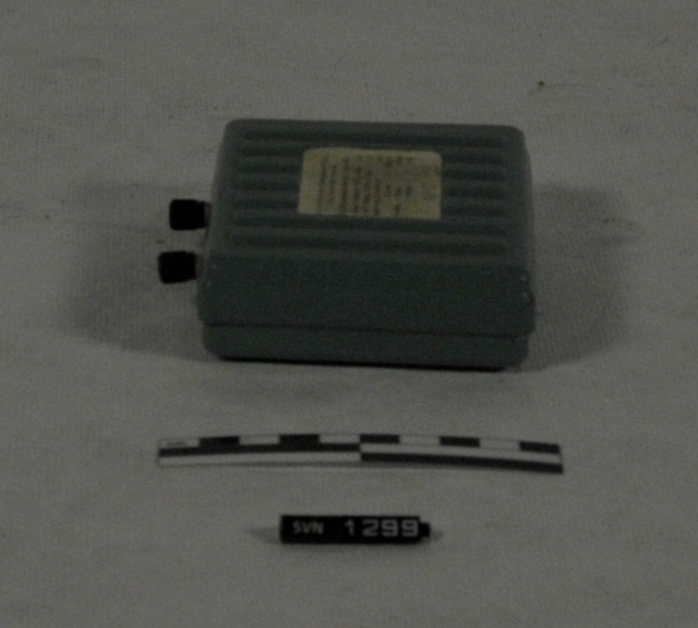 Grå boks med to terminaler for tilkobling av ledninger. Boksen har en hvit papirlapp med spesifikasjoner på oversiden og en sort lapp på undersiden.
