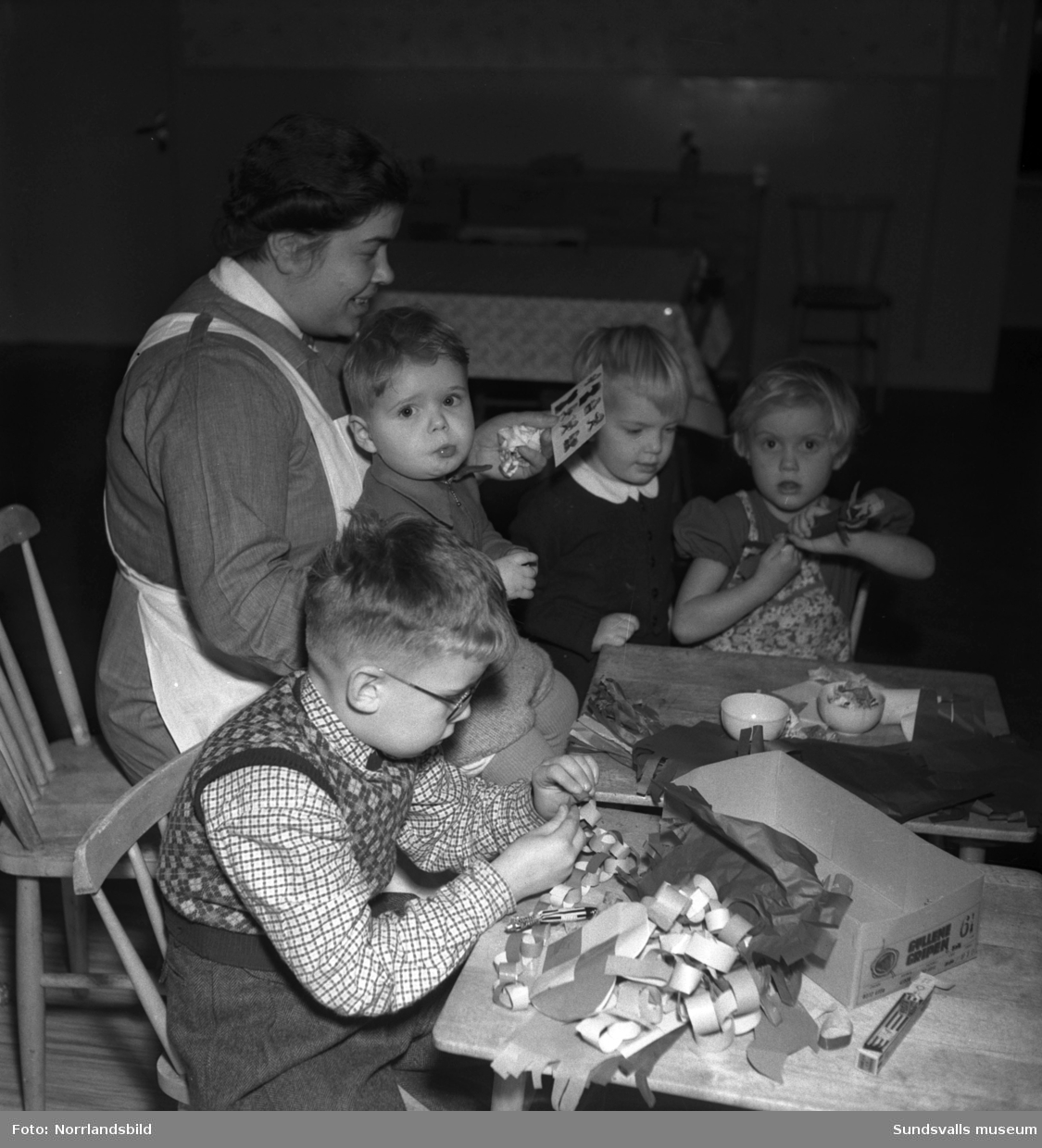 Barn och personal på barnhemmet Morgonsol tillverkar juldekorationer. Branta vägen 8, Södermalm. En liten pojke visar stolt upp en pappersgirlang och ett flätat hjärta.