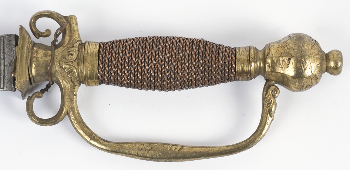 Messing feste fra en kårde. Rund fasettert knapp med nitteknapp.
Tregrep med snodd kobbertråde bevikling og bånd oppe og nede. Fremre parerstang brukket Bakre parerstang går over i håndbøylen, som er tappet inn i knappen.Parerplaten er fjernet, rektangulært baisstykke.
Rett enegget klinge med kileformet tverrsnitt og to smale ryggstillt blodrender.
Klingen har etset dekor med nanvgitte figurer og andre jaktscener.