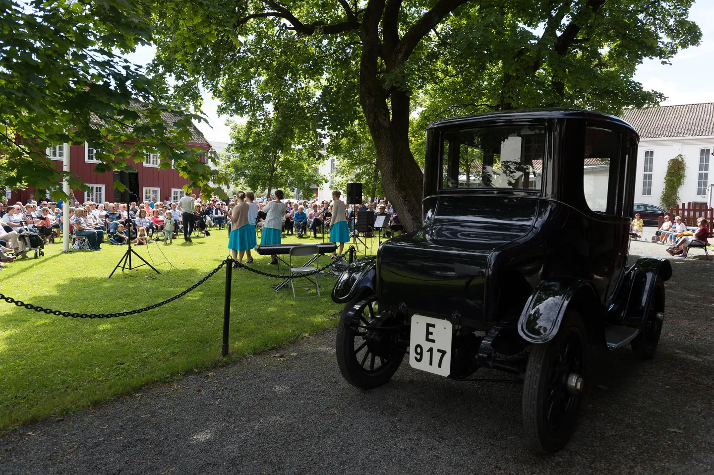 Alf Mjøens Elbil Ohio Electric i forgrunnen. Artister og publikum på tunet på Gjøvik gård. Sol og grønn plen.