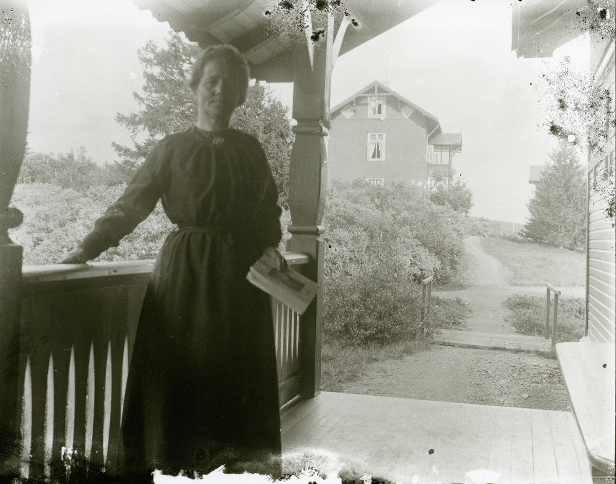 Kvinne som står på en veranda med ei bok i handa.