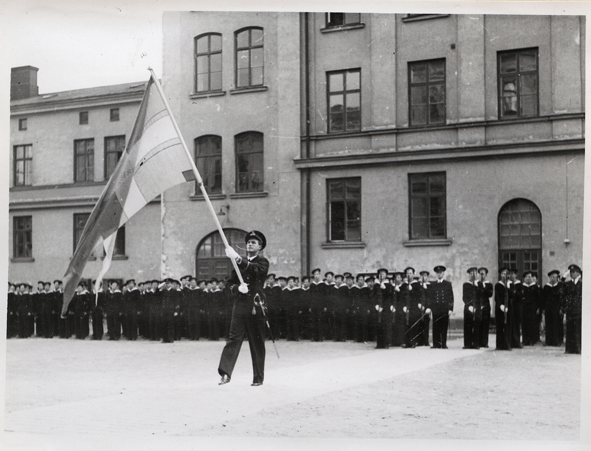 Fotot visar en militär ceremoni på kaserngården i Karlskrona. Centralt i bilden marcherar en fänrik tillika fanbärare med en tretungad örlogsflagga. Han bär mässdräkt komplett med skärmmössa, handskar och också en sabel på vänstra höften. Han håller huvudet högt och hans ansikte ses ur trekvartsprofil. 
En bit bakom honom står yngre män med gevär i händerna, samt två högre befäl varav den ena gör honnör. Bakom dessa står sedan ytterligare yngre män givakt längsmed den kasernbyggnad som utgör fonden.