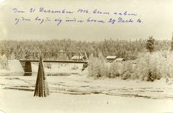 Plassen bru. Tømmer i forgrunnen. Hus i bakgrunnen. Vinter. 