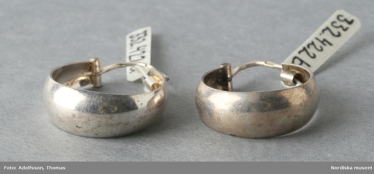 Ett par örhängen, av silver. Breda ringar med låsanordning bestående av smal böjd ten, att fästa genom örat, upptill. Tenen stämplad "925". 
/Leif Wallin 2015-06-08