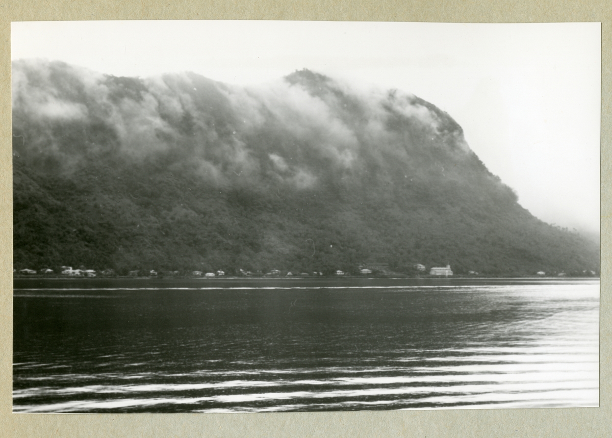 Bilden föreställer ett landskap med vatten och ett trädbeklätt berg. Bilden är tagen under minfartyget Älvsnabbens långresa 1966-1967.