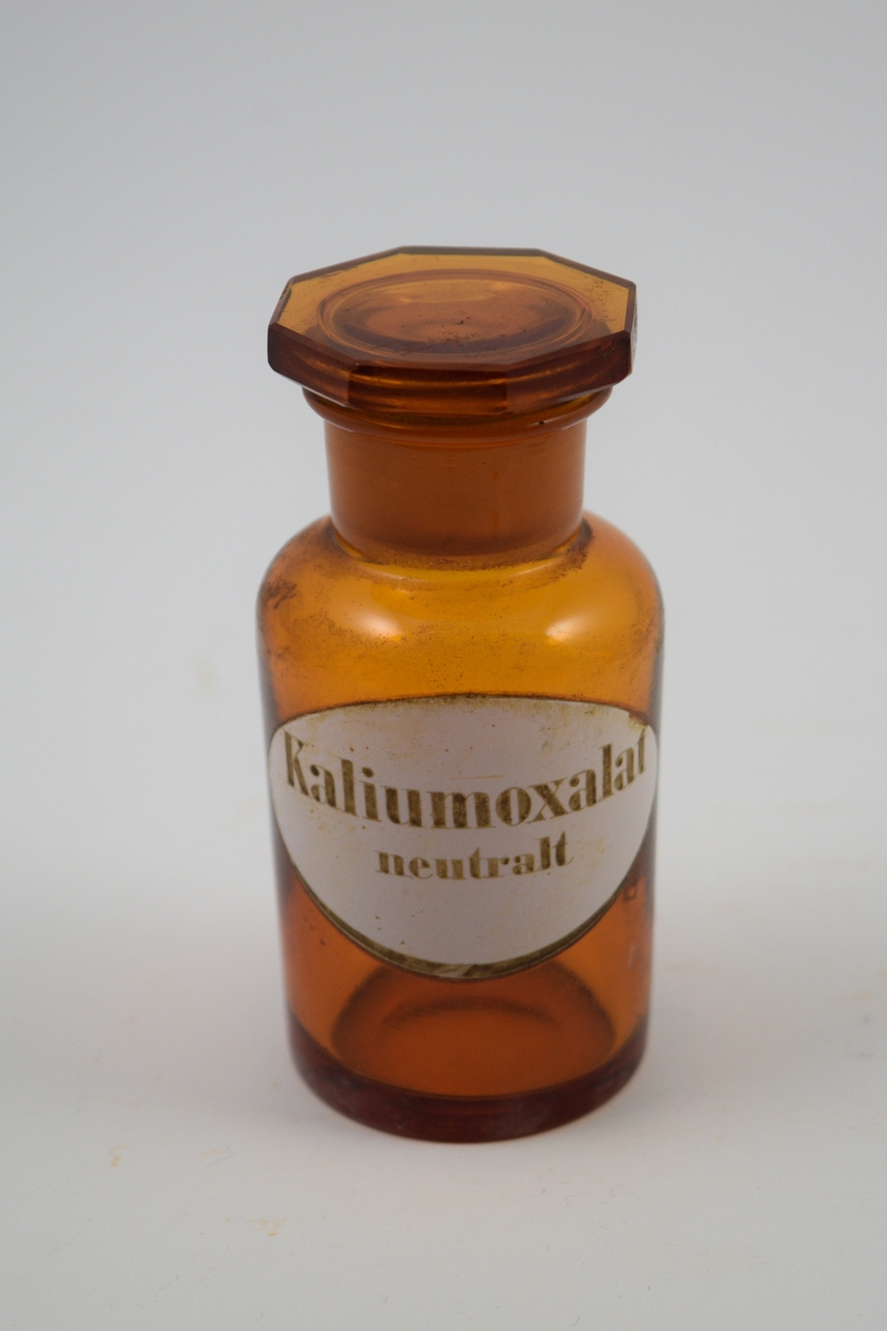 Brun glasskrukke med glasspropp. Etikett hvit med sort skrift. Kaliumoxalat - til bruk i legemiddelproduksjon.