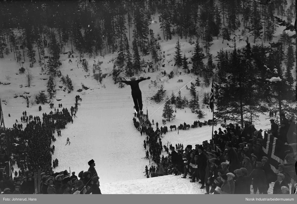Mann hopper på ski i Dalsbakken (mellom Eikeskar og Dalsåsen). Publikum står tett sammen nedover hele bakken, noen med norske flagg.