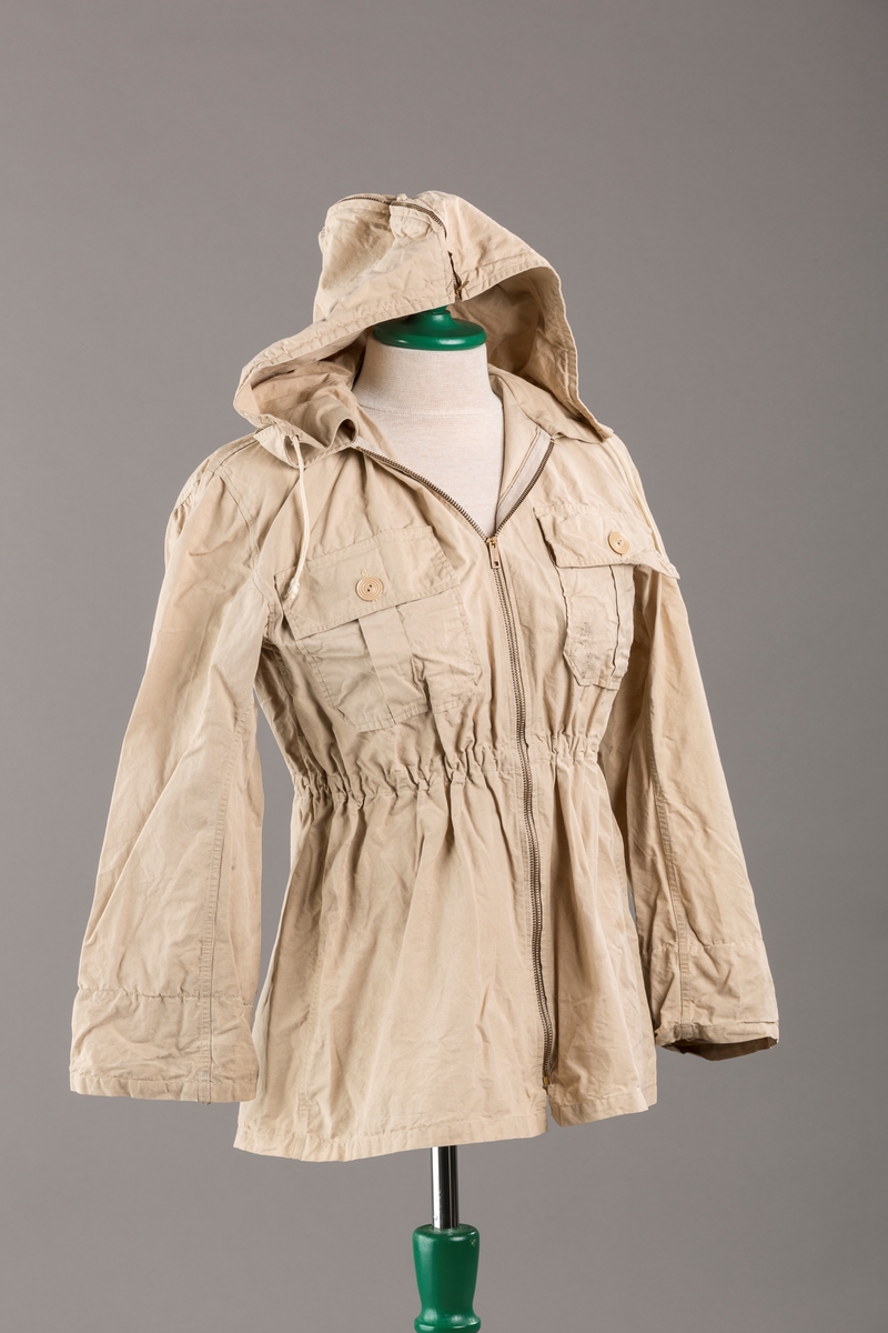 Form: Vanlig anorakkfasong med strikk i livet. 2 brystlommer med knapp.
Hette med glidelås på topp midt, snøring.
Glidelås helt ned foran.
Innermansjett med strikk i ermene.