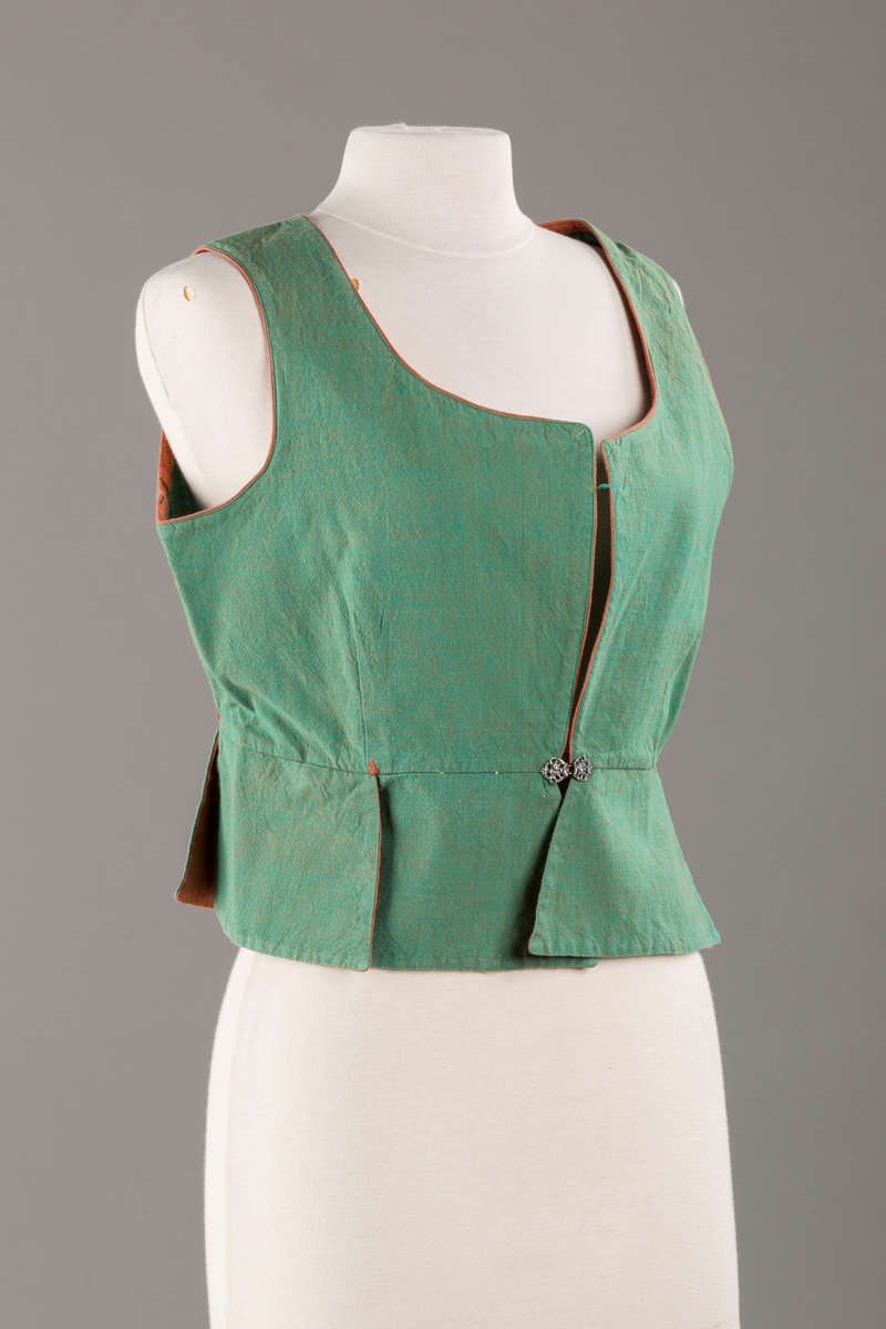 Grønt liv med rustrød kant og fór, seks skjøter. Håndsydd forsterkning i kontrastfarge øverst i skjøtesplittene. Lukkes med tinnhekte.