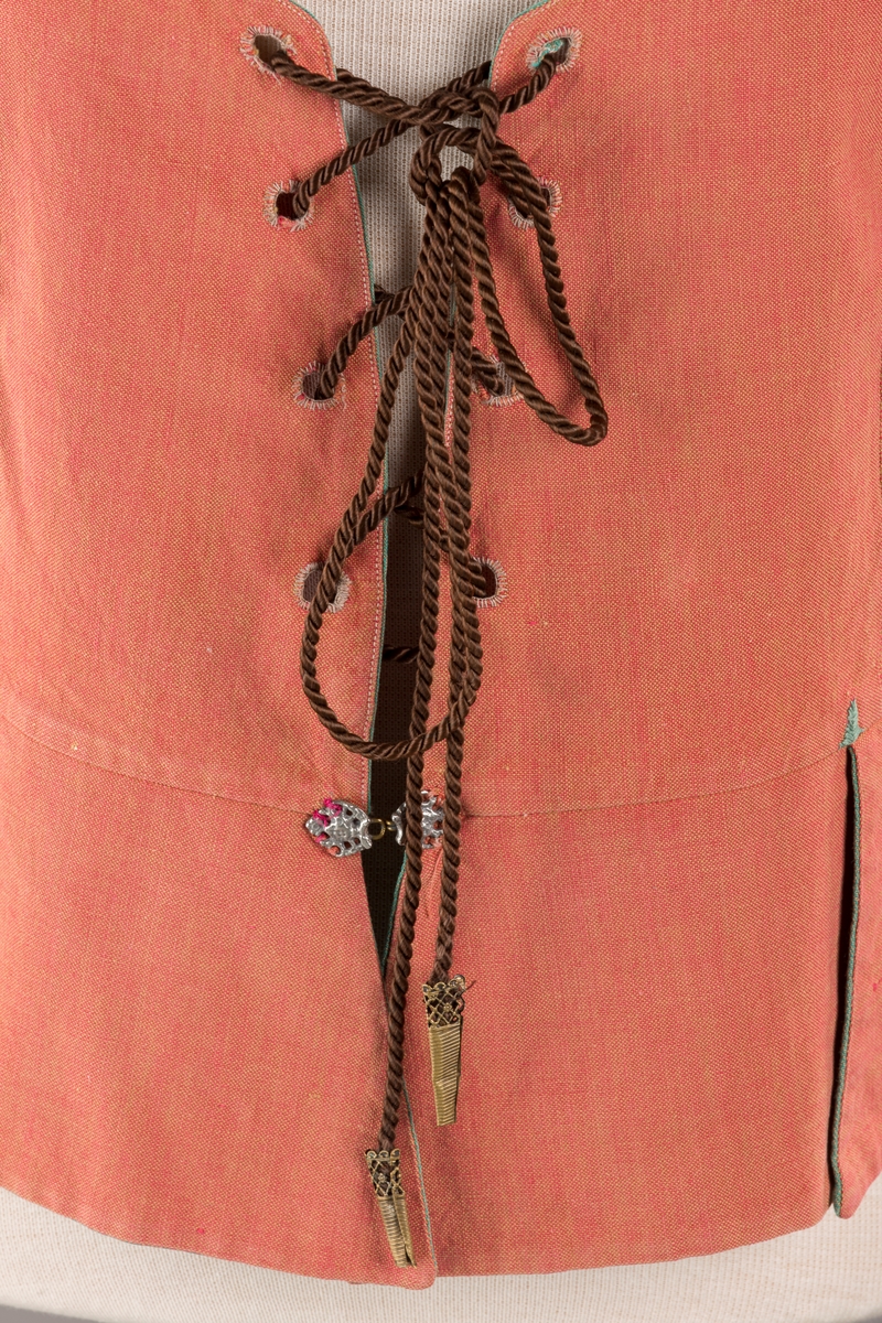 Rustrødt liv med grønn kant og fór, seks skjøter. Håndsydd forsterkning i kontrastfarge øverst i skjøtesplittene. Lukkes med tinnhekte og silkesnor.