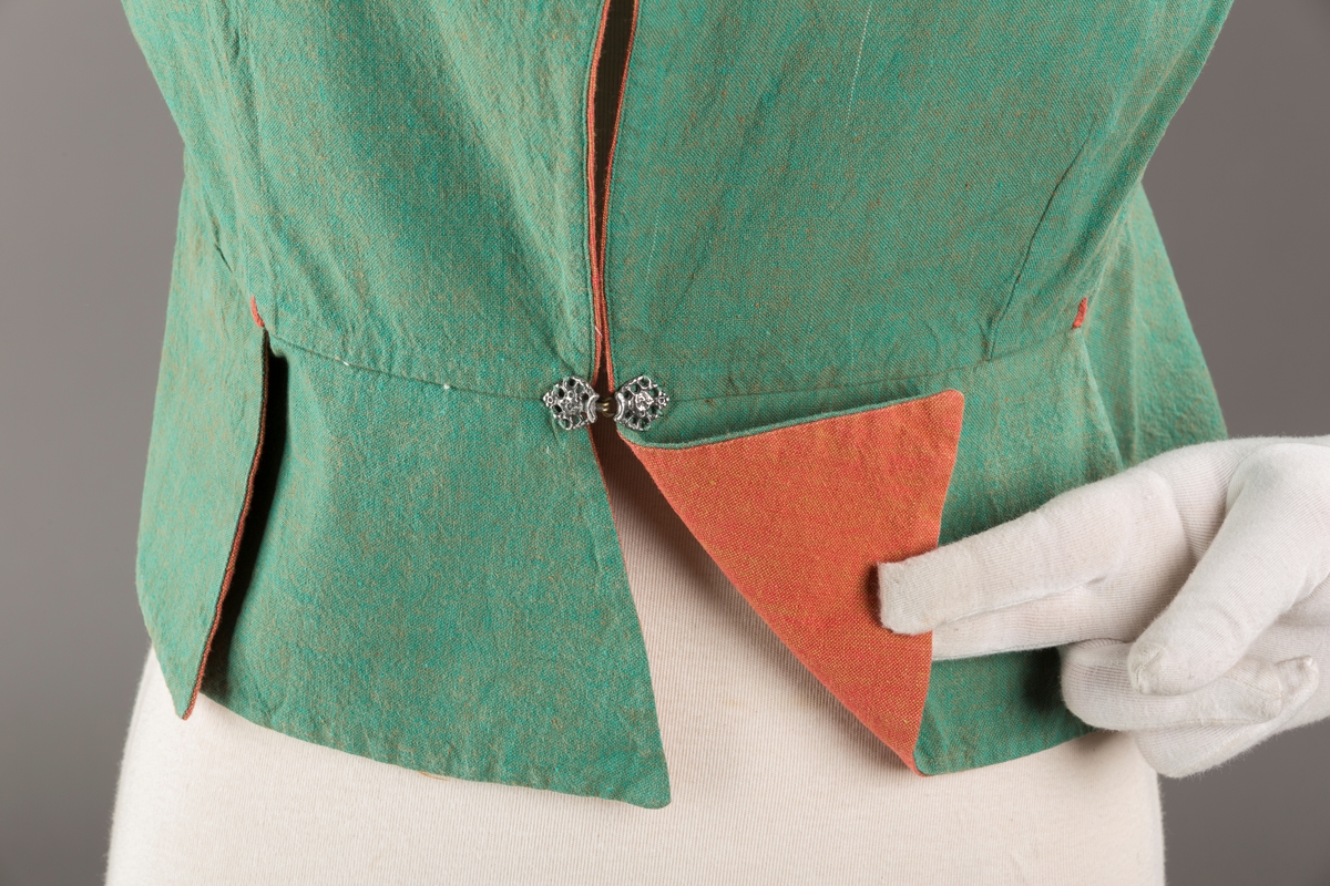 Grønt liv med rustrød kant og fór, seks skjøter. Håndsydd forsterkning i kontrastfarge øverst i skjøtesplittene. Lukkes med tinnhekte.