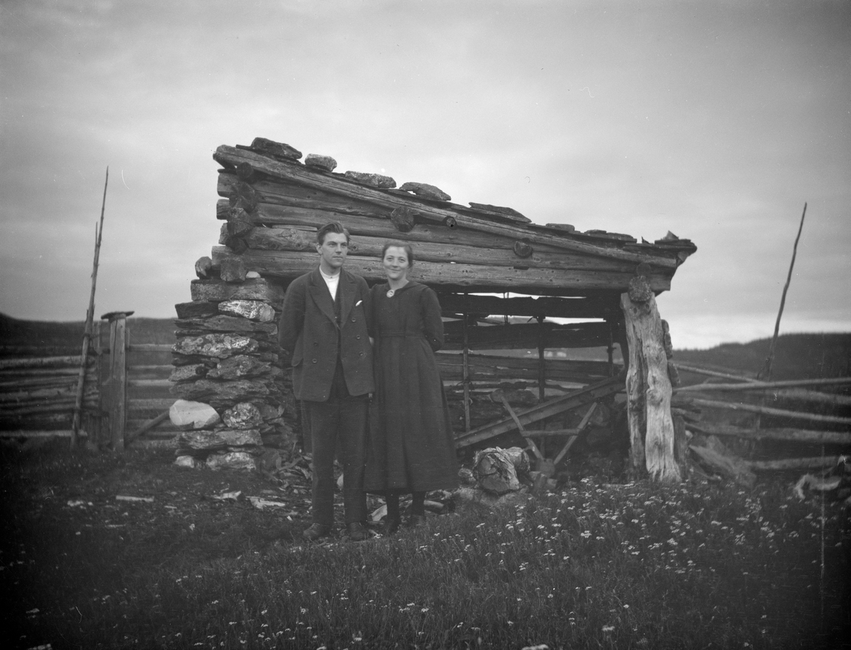 Ungt par står forran et enkelt lavt skur med stein mur på ene siden, to luftige tømmervegger og åpen i front. Ligger steiner på takets forkant.