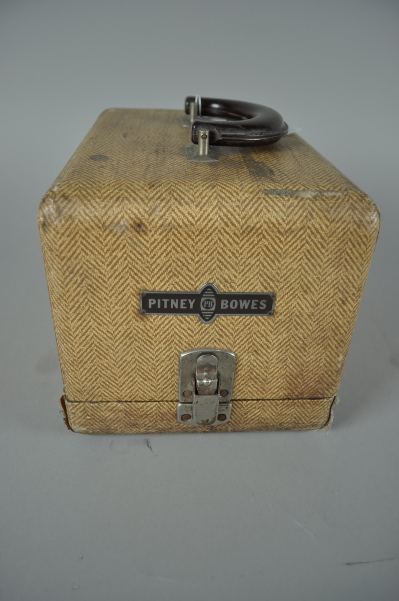 Frankeringsmaskin av brunt metall. 

Maskinen er plassert i trekasse med fiskebensmønstret utside. Kassen har håndtak.