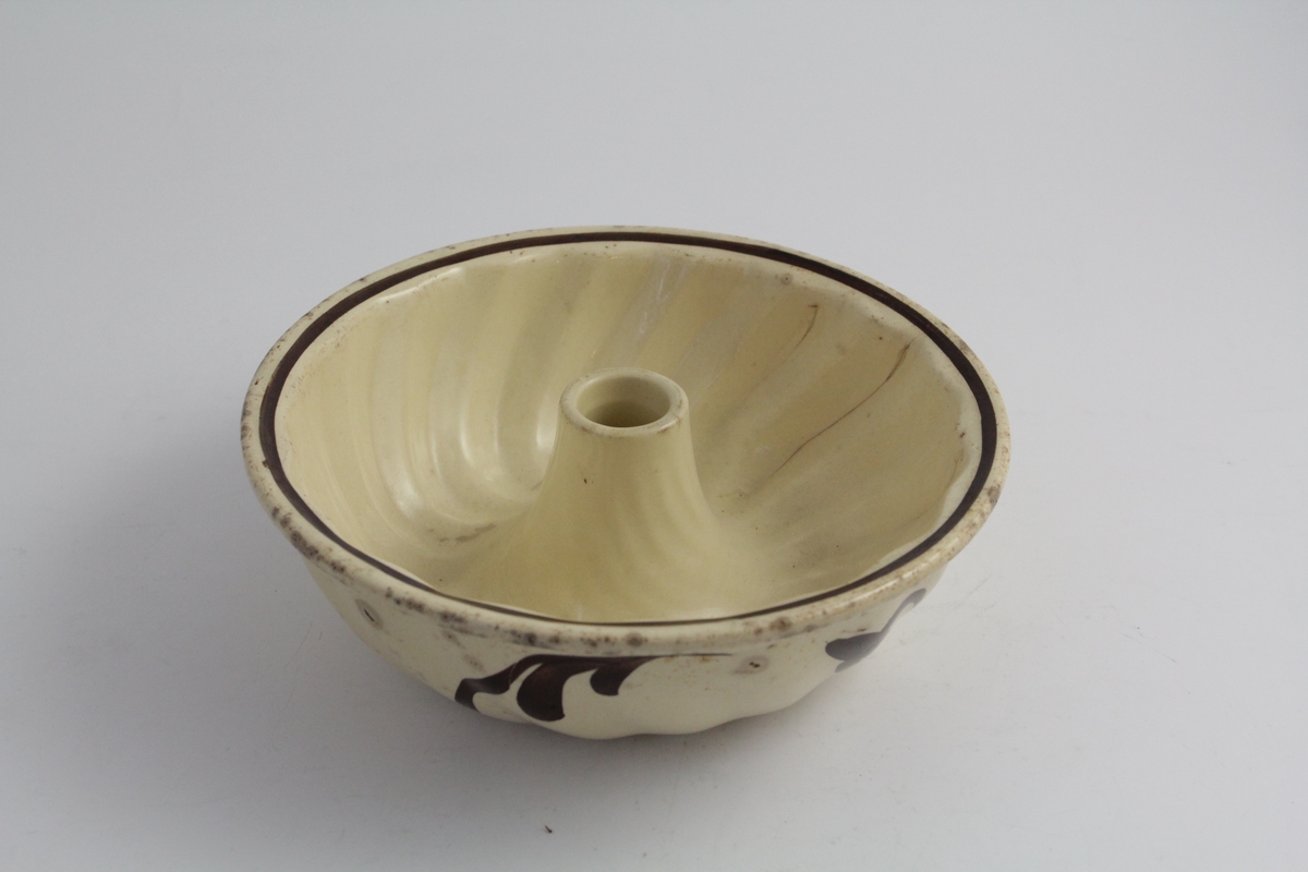 Rund form i ildfast steintøy. Kremgul farge, med brun dekor. Brune stiliserte blomsterkvister på sidene. Rommer halvannen liter.