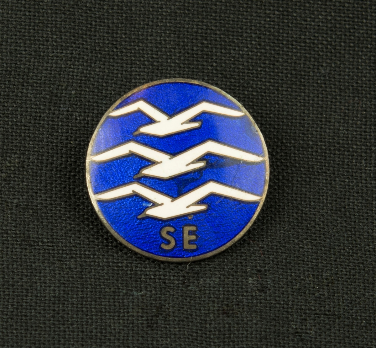 Svenska segelflygförbundets märke i emajl, motivet är tre vita fåglar på blå botten med texten SE i guld. Fästes med nål på baksidan.