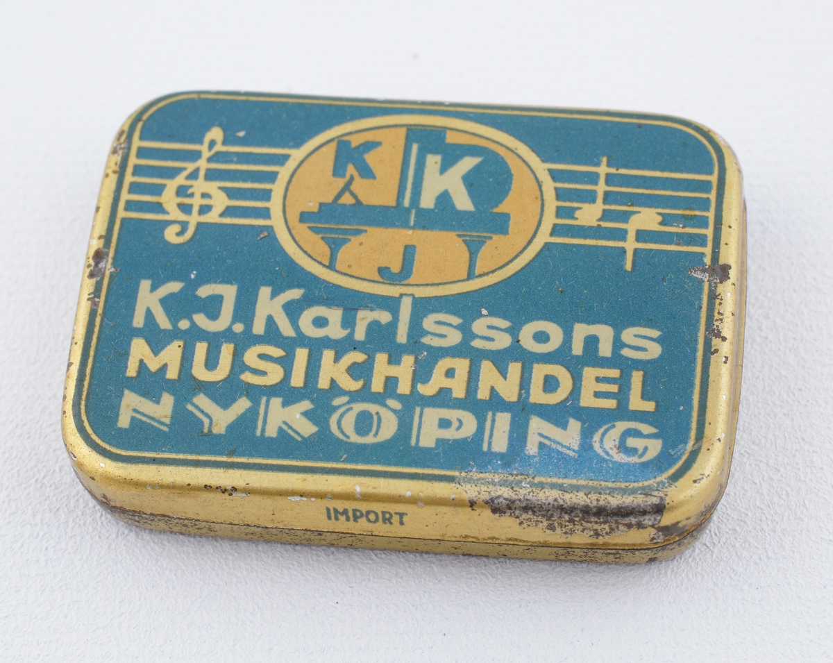 Ask av plåt.
Fyrkantig modell med avrundade hörn och dekor i blått och guld. Förvaringsask för grammofonnålar. 
Text på locket, "K.J. Karlssons Musikhandel Nyköping".