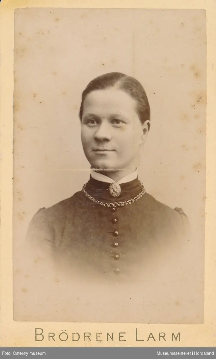 portrettfotografi av ung kvinne med midtskill og håret samla i nakken, mørk kjole med kvit krage og sølje, kjede om halsen