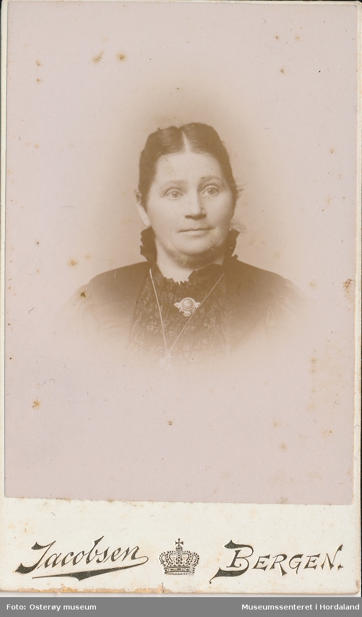 portrettfotografi av kvinne med midtskill, mørkt hår smala i nakken, mørk kjole med brosje og kjede i halsen
